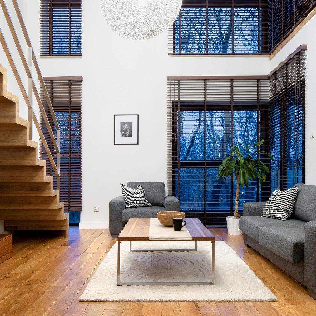A double-height lounge with a timber staircase, grey sofas, a white rug and tall windows with venetian blinds