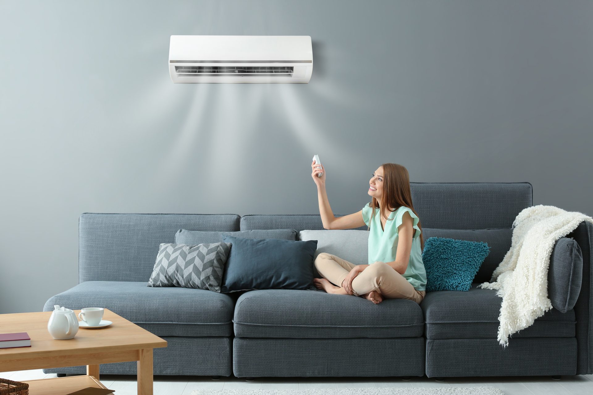 A woman on a grey sofa pointing a remote at a wall-mounted heat pump above her