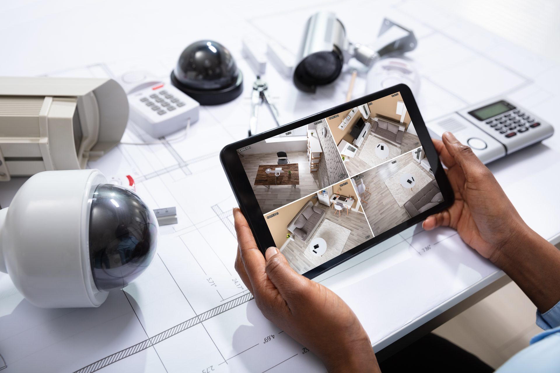 Hands holding a tablet showing four security camera views of a house, with dome and bullet cameras and an alarm keypad on the desk